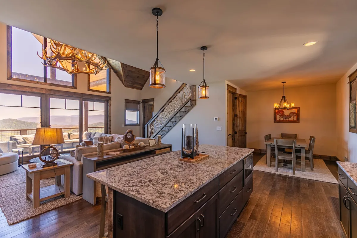 This image showcases a spacious open-concept interior where a rustic chandelier hangs over a wooden dining table and a granite-topped kitchen island. Large windows flood the room with natural light and frame scenic mountain views, while the space flows easily between kitchen, living, and dining zones.