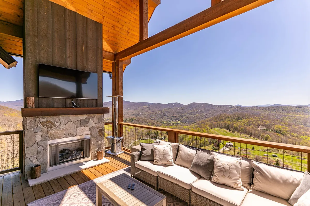 An elegant outdoor seating area features plush chairs arranged around a contemporary stone fireplace. The space is open to the mountain landscape and includes a television, blending outdoor comfort with entertainment. The view extends to the mountains, offering a picturesque setting for al fresco dining or cozy gatherings.