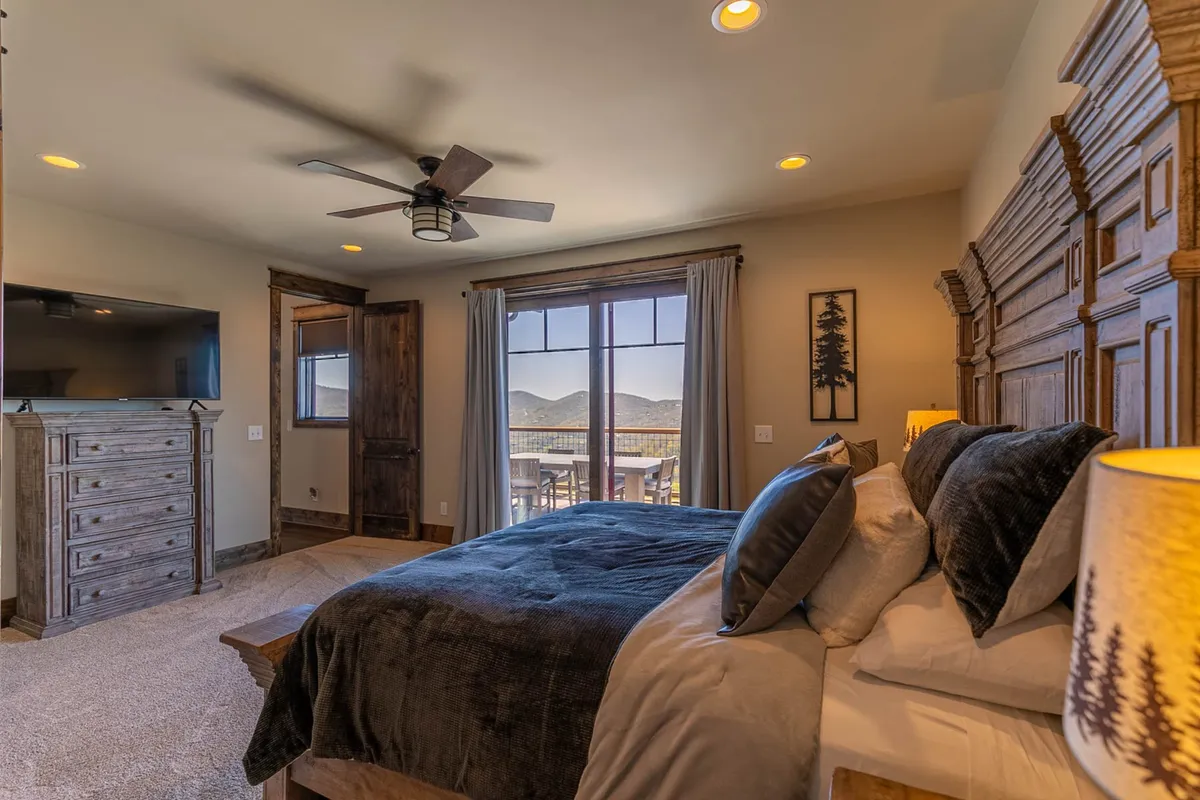 This serene bedroom features a plush king-size bed, a wooden dresser, ceiling fan, and a large window that frames stunning mountain views. The tranquil atmosphere is perfect for unwinding after a day exploring the outdoors.