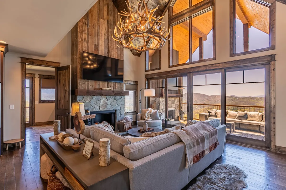 A cozy sectional sofa is arranged around a striking stone fireplace, with an antler chandelier above and expansive windows bringing in mountain vistas. The scene is set for comfortable relaxation or lively gatherings with family and friends.