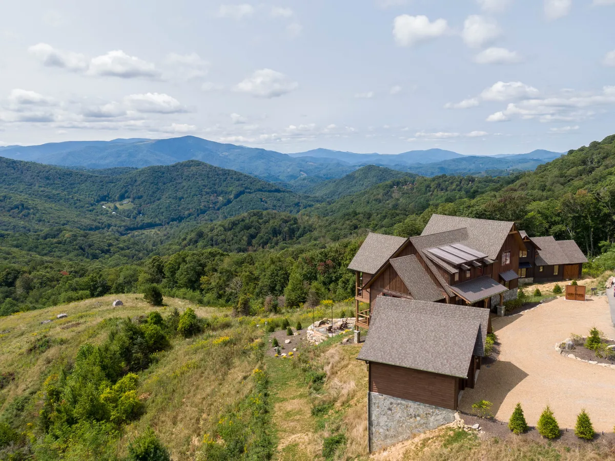 The image showcases Bear Haven Hideaway, a stunning mountain home set against a dramatic Blue Ridge backdrop. The beautifully landscaped garden frames the house, while the spacious porch offers sweeping sunrise views. An inviting outdoor fire pit hints at cozy evenings under the stars, all surrounded by tranquil woods and the serenity of nature.
