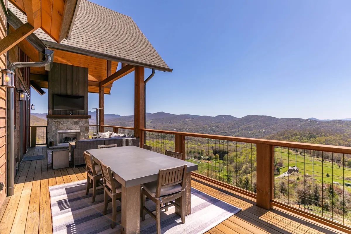 This image showcases the main level deck with a spacious outdoor dining table, a cozy sectional seating area, a modern gas fireplace, and a mounted TV, all set against a beautiful panoramic mountain view. The scene captures the area set up for social gatherings and relaxing, with natural light highlighting the inviting atmosphere.