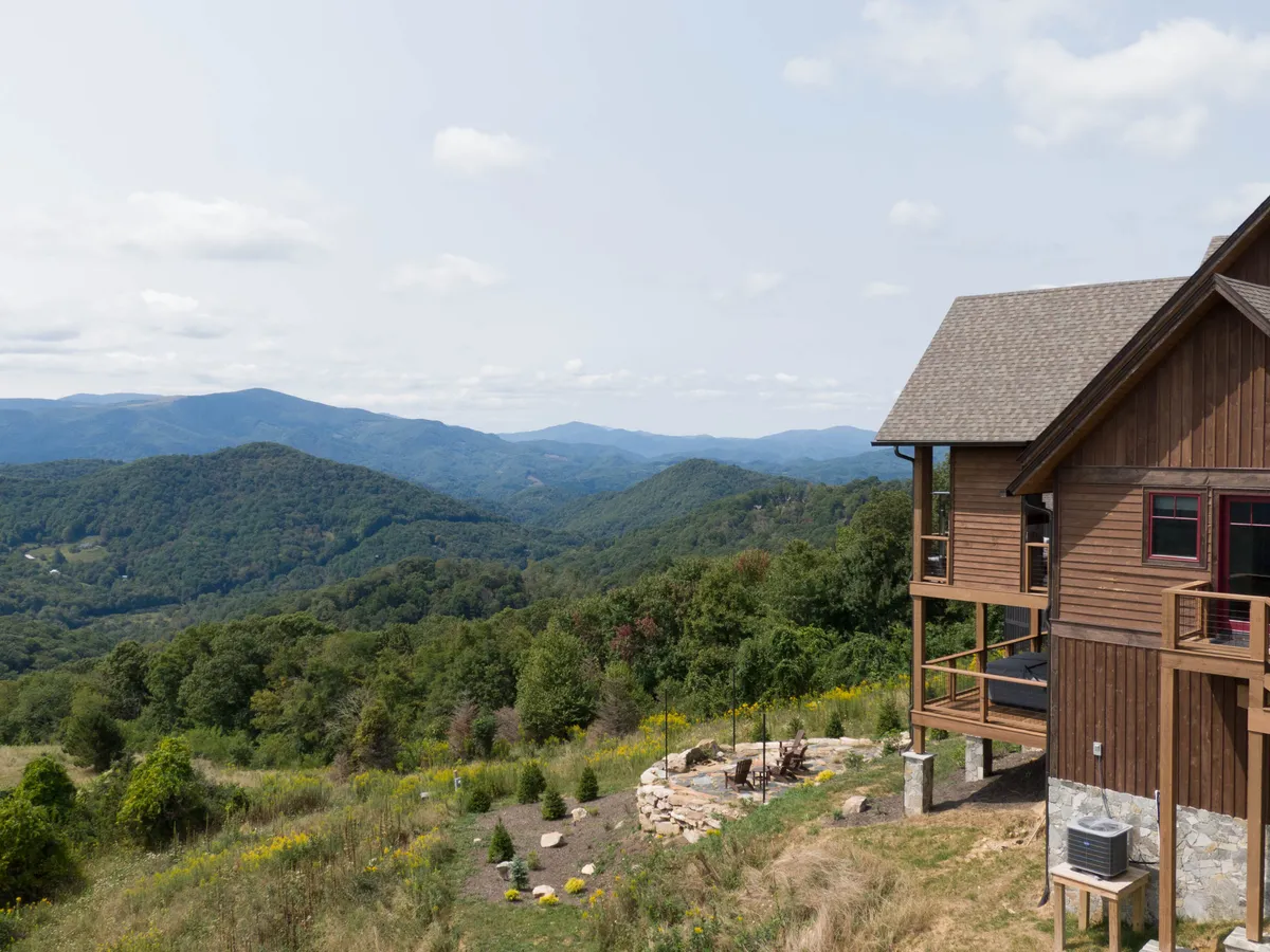 This inviting exterior image features a classic wood-burning fire pit surrounded by rustic chairs, with a backdrop of lush greenery and the majestic mountain range. The wooden house is visible, enhancing the cozy, communal atmosphere.