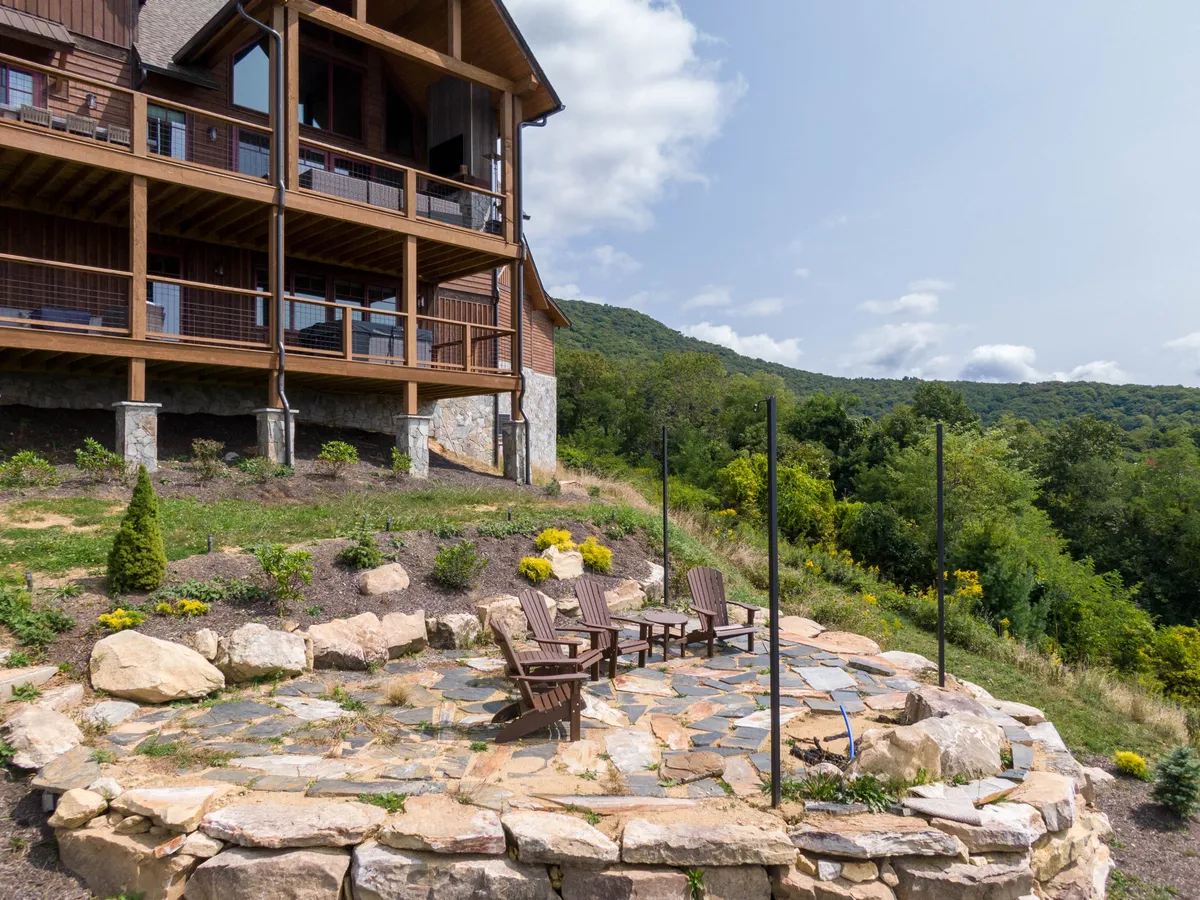 This image captures a tranquil stone patio with Adirondack chairs facing a landscaped garden and a wooden balcony, all set against a lush natural backdrop. The space is perfect for morning coffee, evening bonfires, or simply enjoying the fresh mountain air and sunset views.