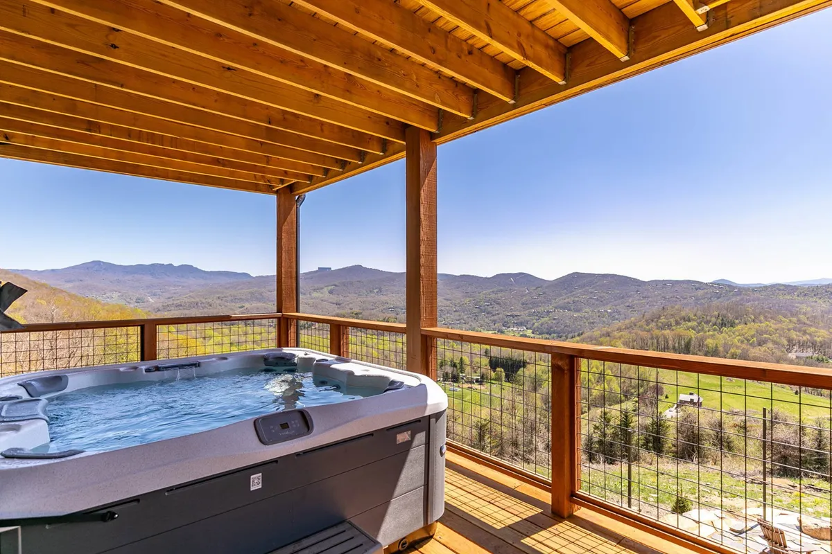 This exterior image features a luxurious hot tub set on a wooden deck, framed by sturdy wooden beams, with expansive views of the surrounding mountains. The tranquil ambiance is accentuated by the serene mountain backdrop, inviting guests to relax and socialize in a resort-style setting.