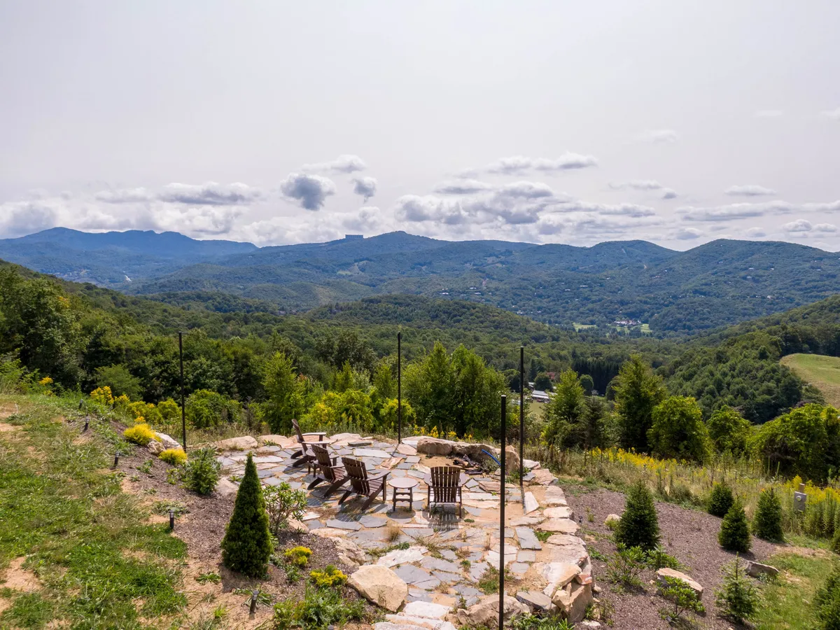 This view image emphasizes the breathtaking mountain landscape surrounding Bear Haven Hideaway, with a stone patio and wooden chairs positioned perfectly for admiring the panoramic scene and lush greenery. The image evokes peaceful reflection and the allure of nature in all seasons.