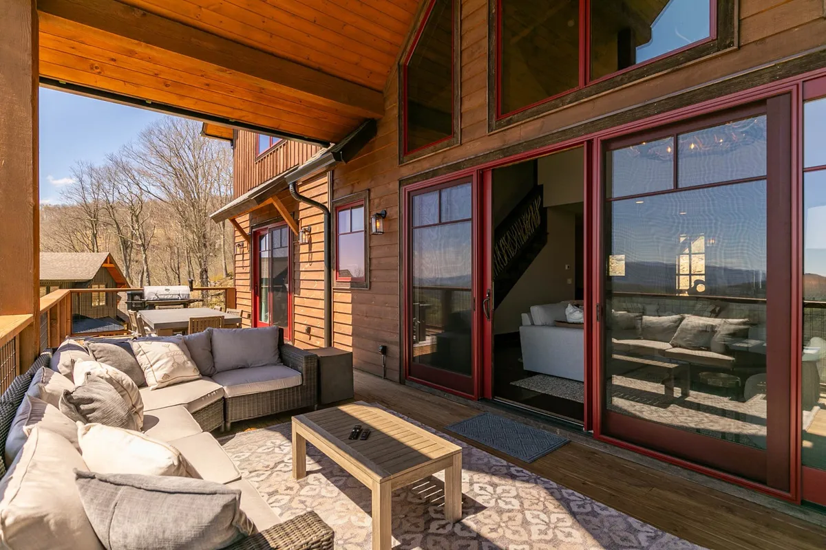 This image shows the inviting exterior of Bear Haven Hideaway, featuring a comfortable outdoor seating area, large windows, and charming wooden accents. The patio and seating area are designed for relaxation, social gatherings, and soaking in the mountain atmosphere, with hints of nature visible around the home.