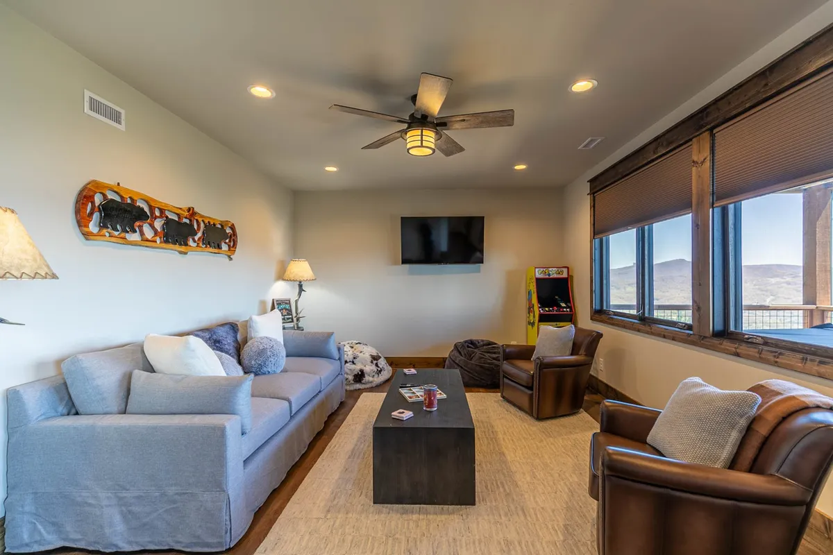 This image displays a casual and comfortable den featuring a sofa, coffee table, wall art, a large television, and an arcade game machine. The layout is open and inviting, with the arcade machine prominently visible beside the TV, highlighting the room’s entertainment focus.