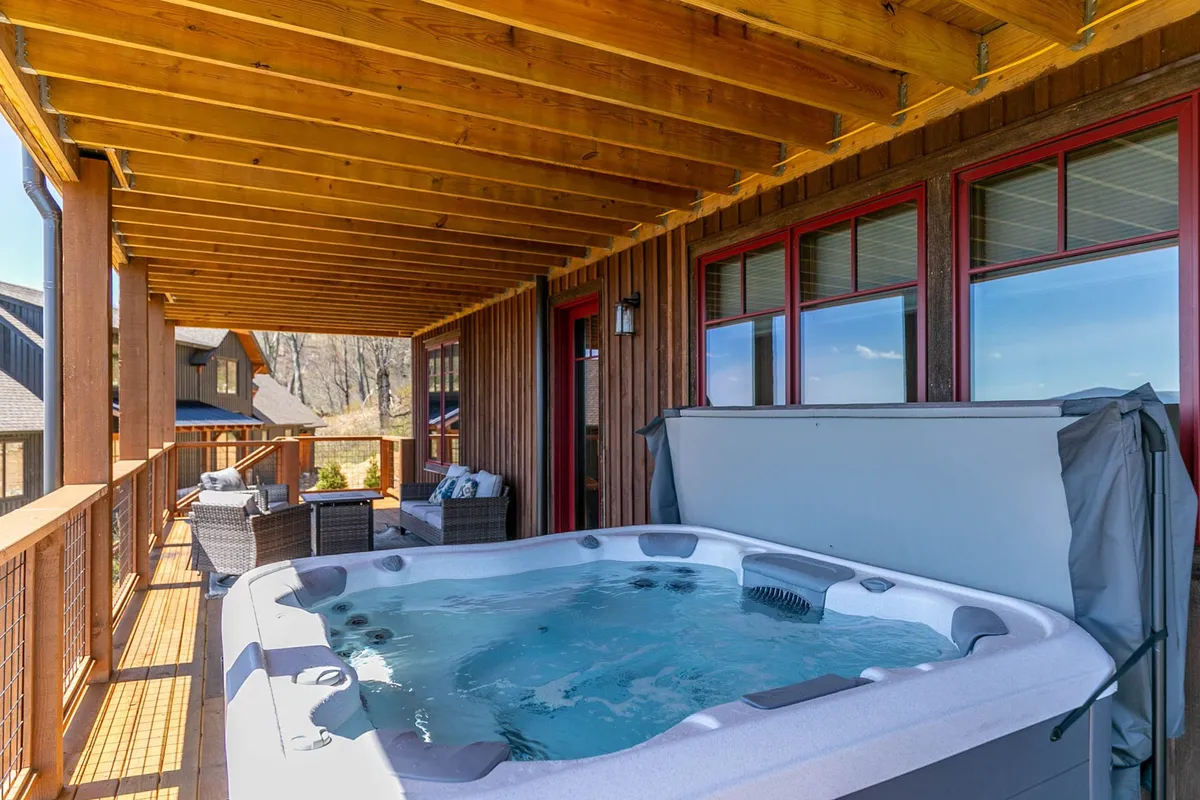 An inviting exterior deck features a hot tub surrounded by wooden beams and windows framed in red trim. The seating area and lush scenery offer a tranquil spot to unwind, ideal for stargazing after a day of mountain adventures.